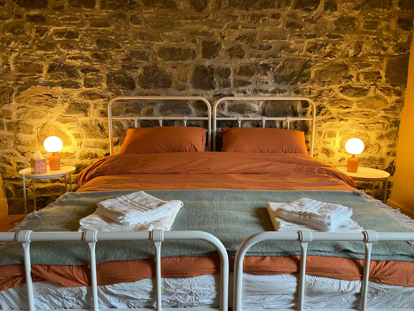 Double bedroom at night: iron beds, exposed stone wall lit by warm bedside lamps, terracotta linen and folded towels