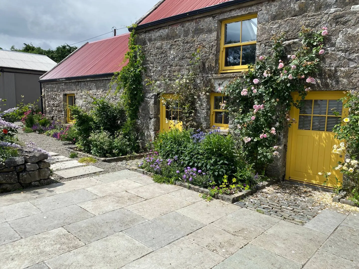 Farmhouse front in full summer bloom: climbing roses, yellow doors and windows, stone walls, a path through the flowers