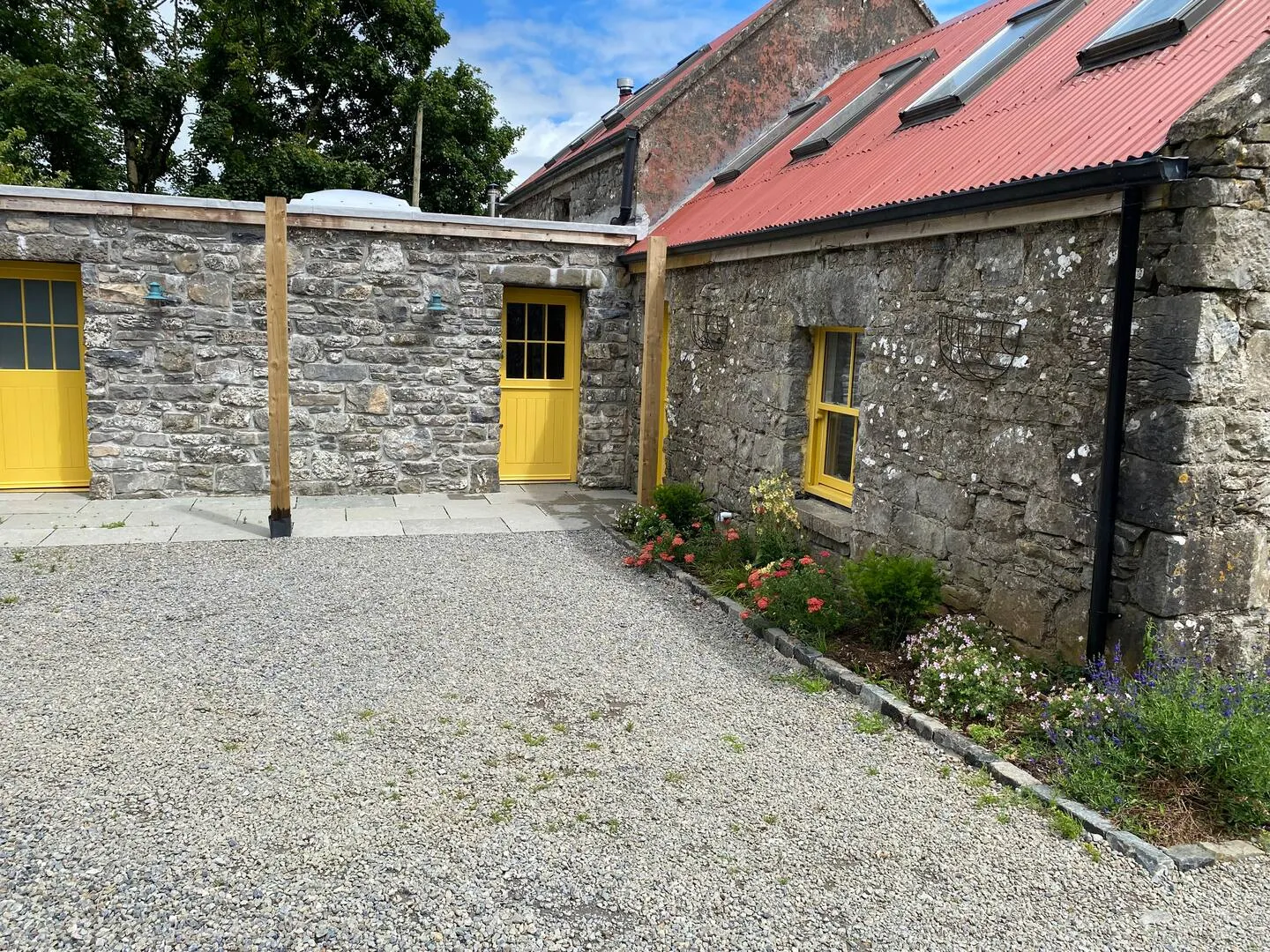 Stables courtyard from the side: yellow door and window, stone walls, red tin roof, gravel and summer flower border