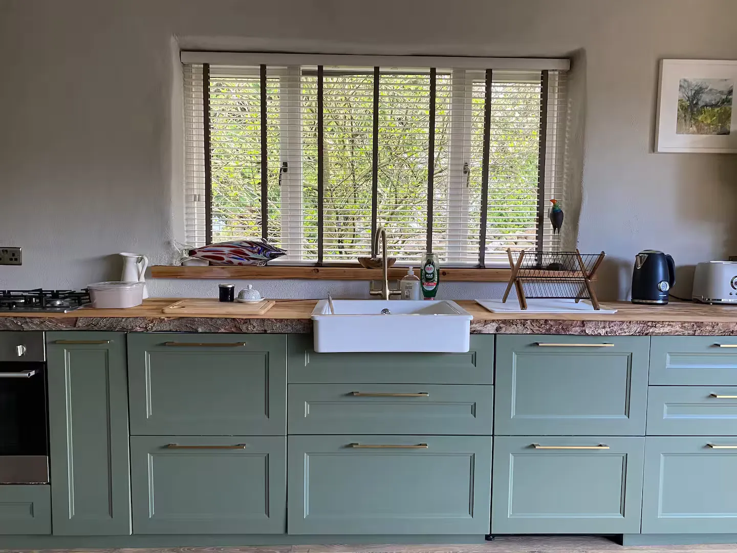 Kitchen close-up: sage green cabinets, Belfast sink, live-edge timber worktop and window to trees