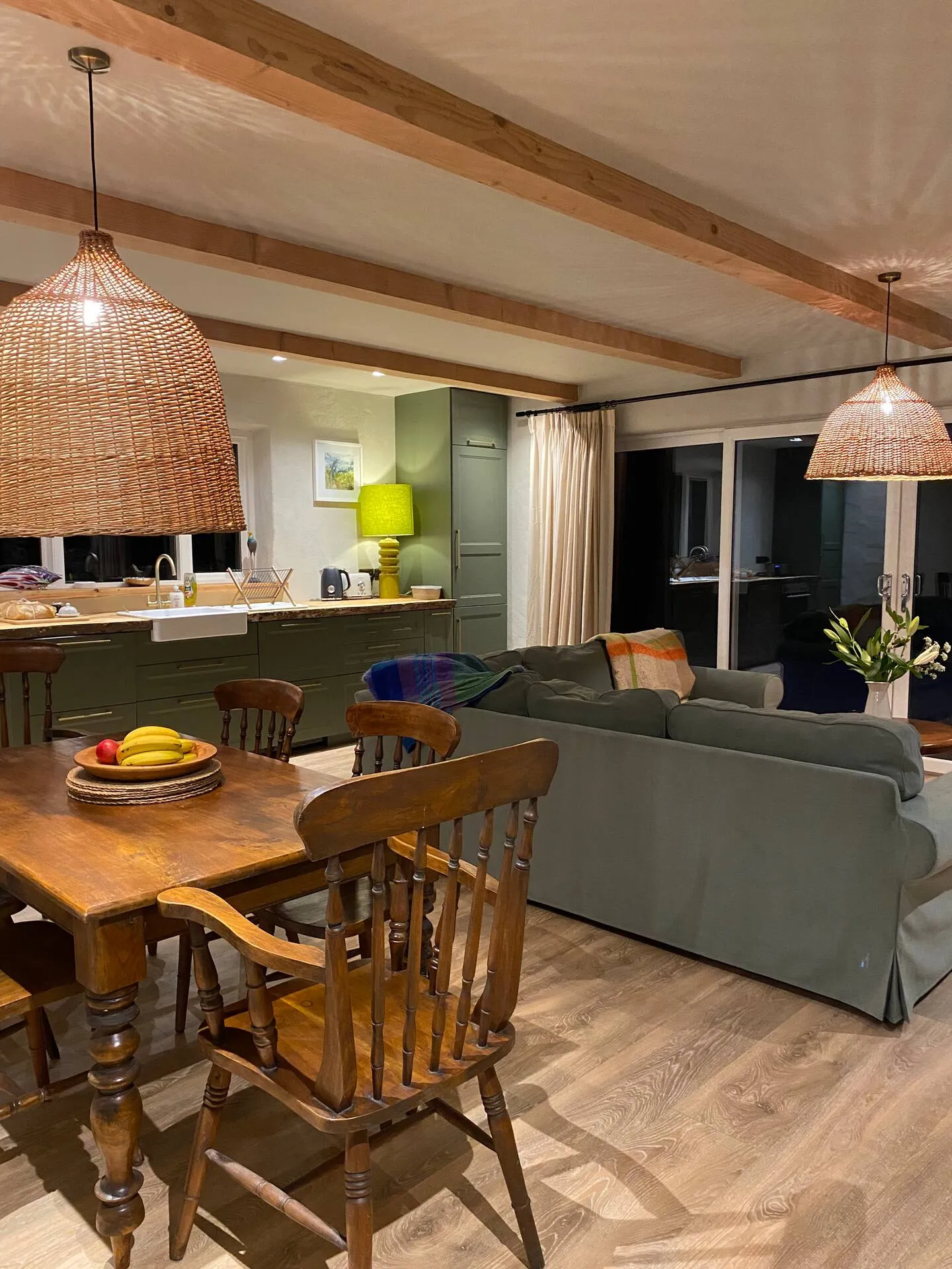 Farmhouse dining table in the evening with large wicker pendant lights, green cabinets and Belfast sink beyond