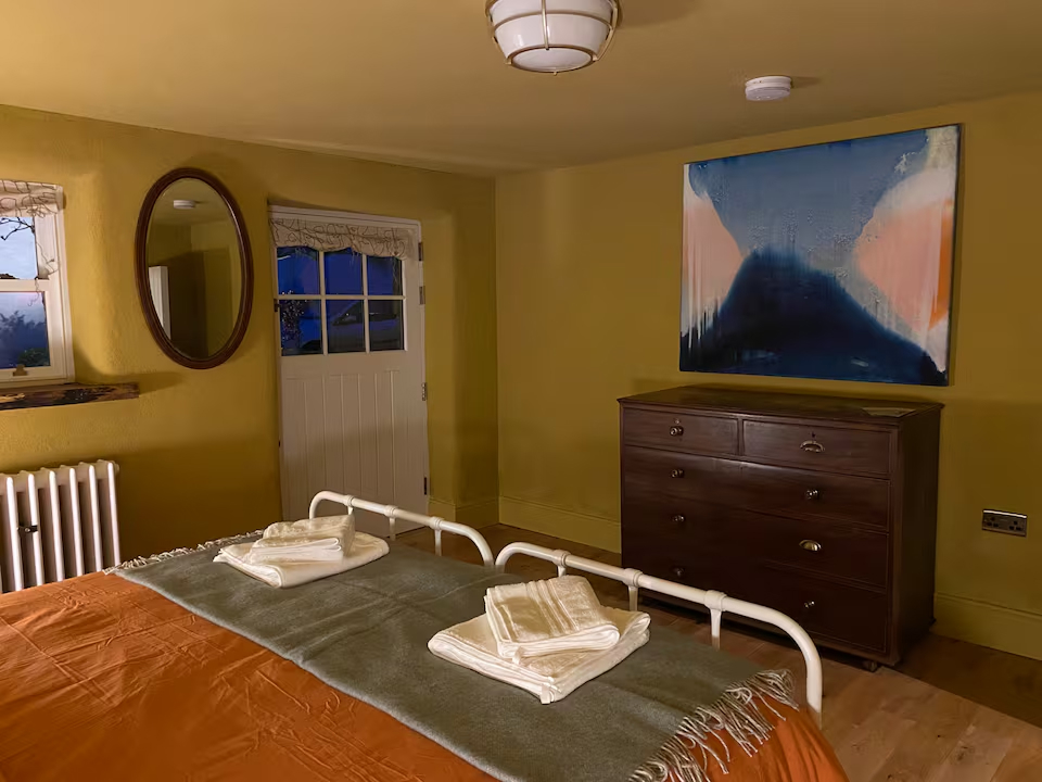 Bedroom with deep mustard walls, large abstract painting, antique chest of drawers and folded towels