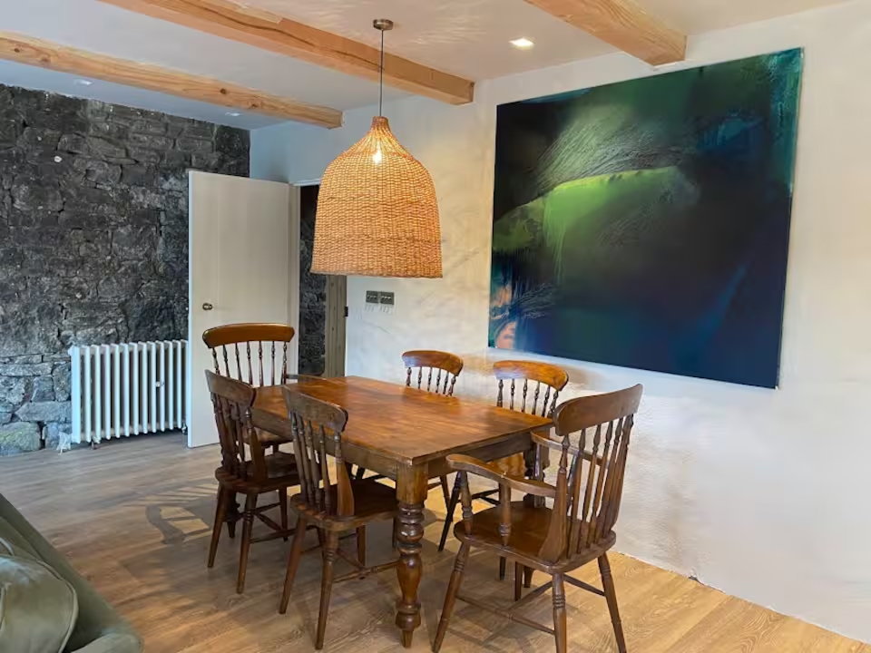 Farmhouse dining table with spindle chairs beneath a large wicker pendant, original stone wall and abstract painting