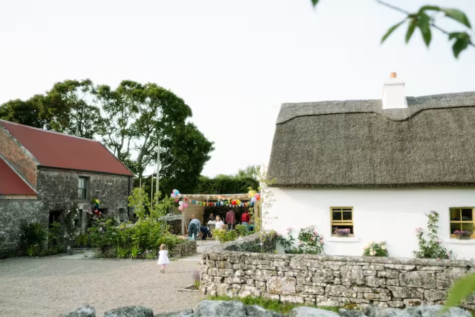 The farmyard with bunting and guests gathered — the thatched cottage and red barn roof visible