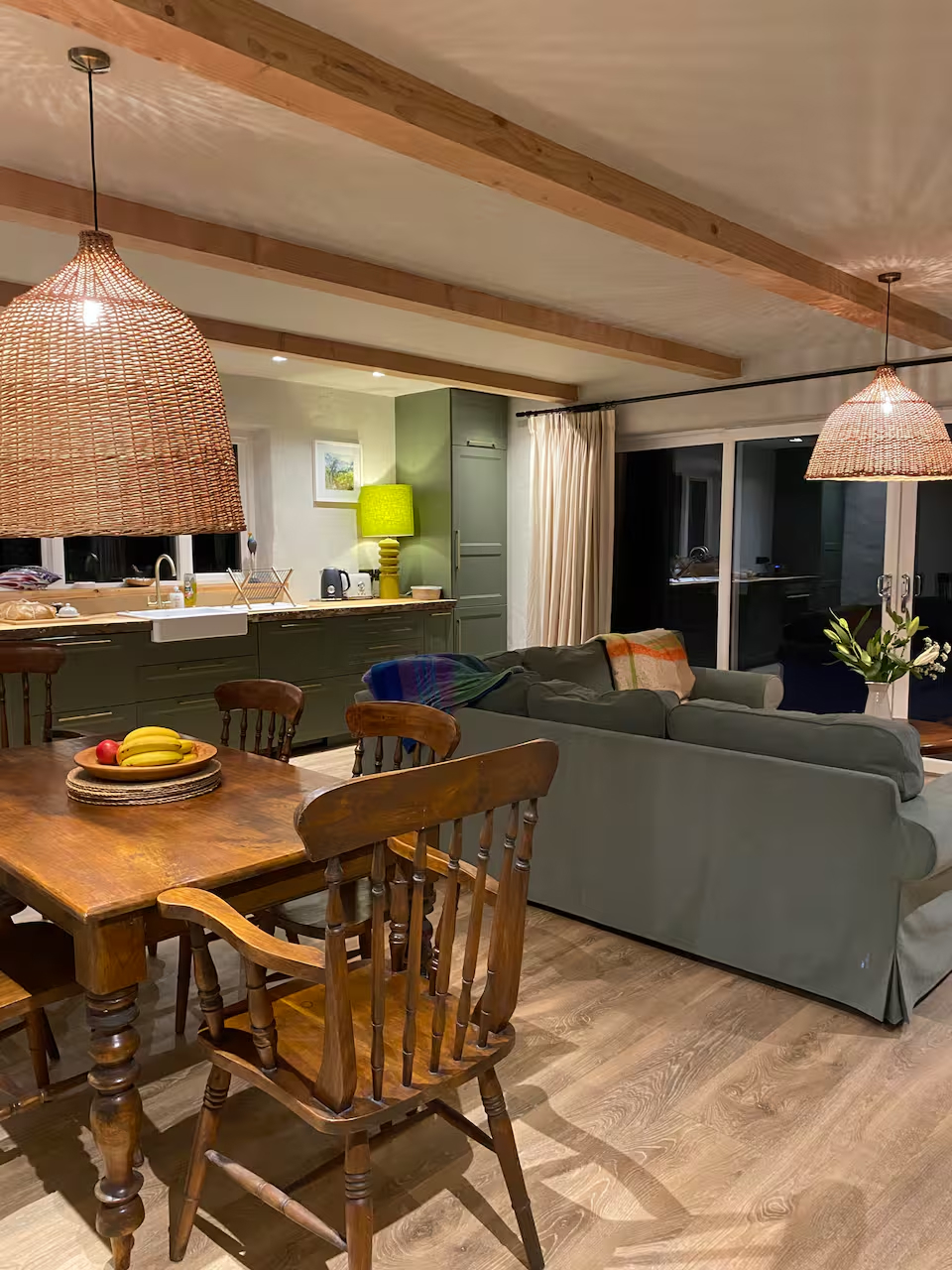 Open-plan living and dining with exposed timber beams, bowl of fruit on the table, kitchen visible